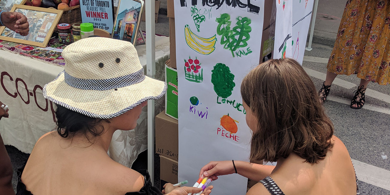 Two women choose markers to draw produce on a mock produce shelf.