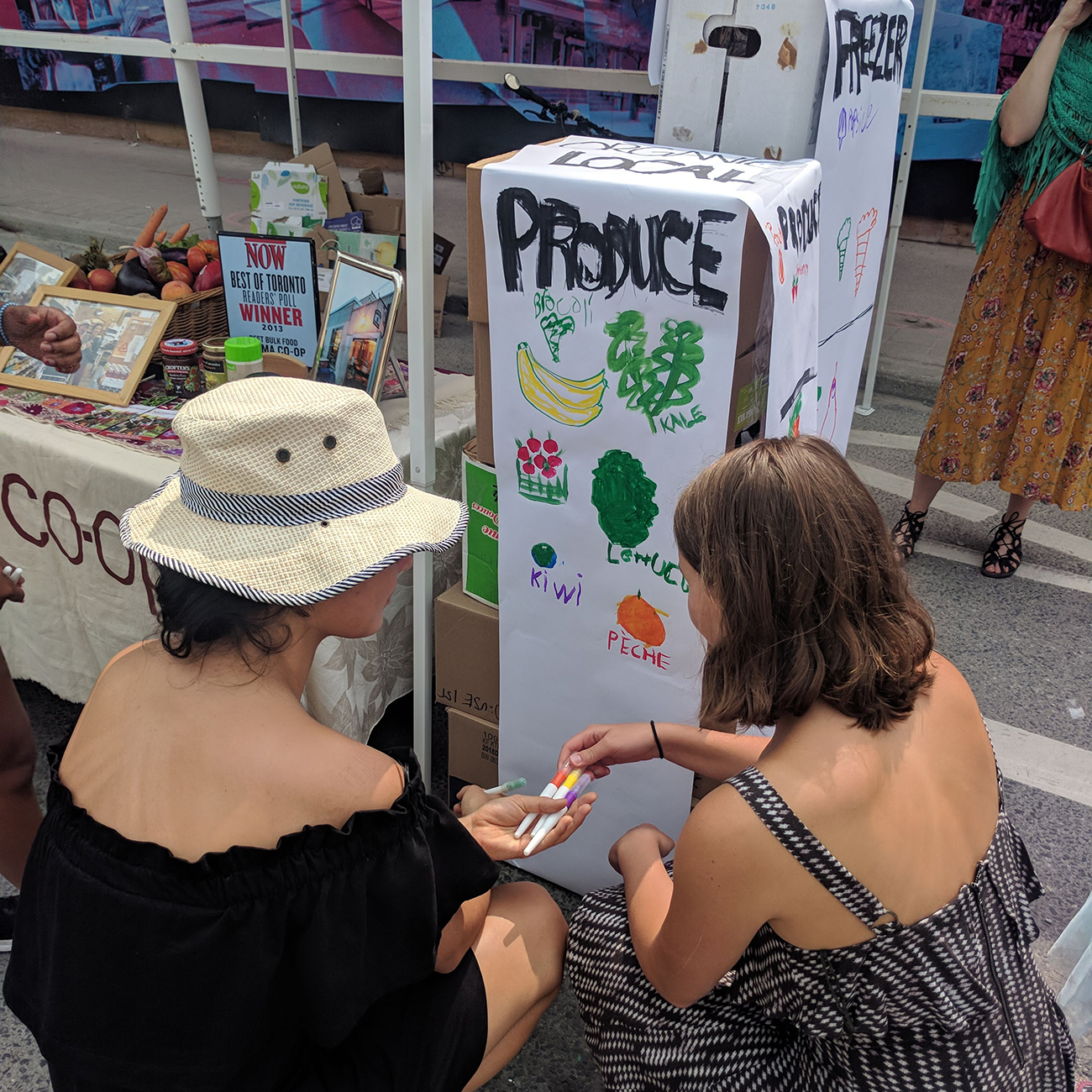 Two women choose markers to draw produce on a mock produce shelf.